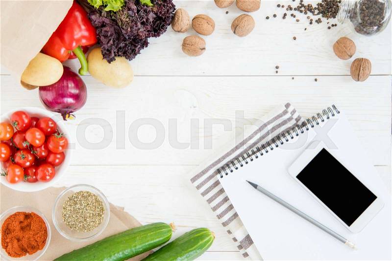 Top view of smartphone with blank screen, empty notebook with pencil, kitchen towel and fresh ingredients on wooden table , stock photo
