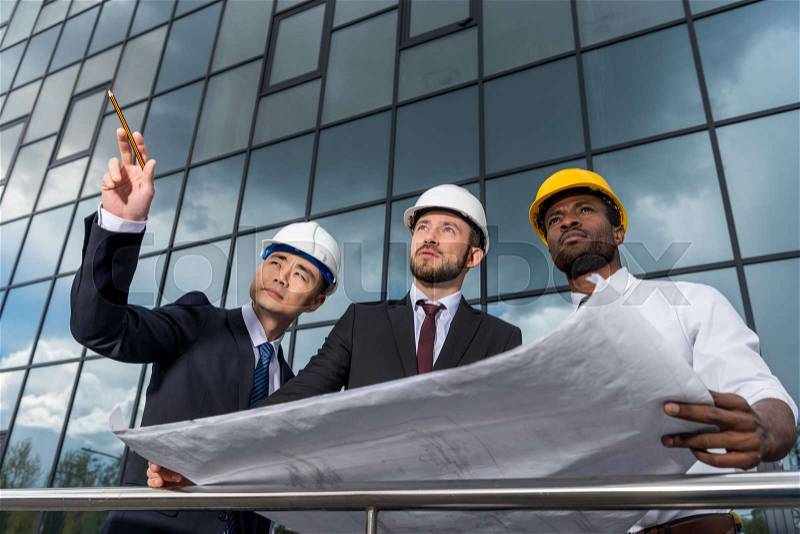 Multiethnic group of professional architects in helmets working with blueprint outside modern building, stock photo