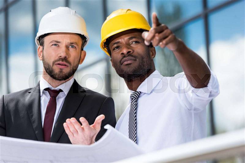 Professional architects in hardhats working with blueprint outside modern building, stock photo
