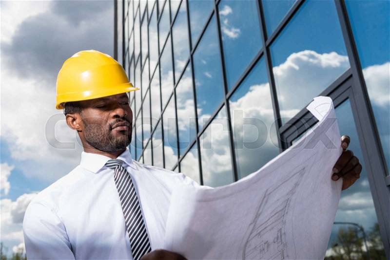 Low angle view of professional architect in hard hat holding blueprint outside modern building, stock photo
