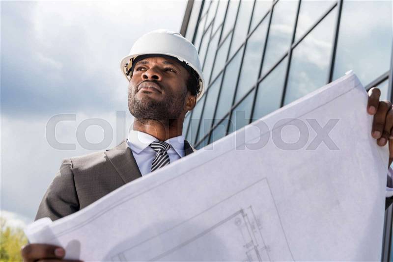 Low angle view of professional architect in hard hat holding blueprint outside modern building, stock photo