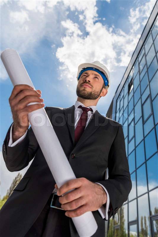 Low angle view of professional architect in hard hat holding blueprint and looking away, stock photo
