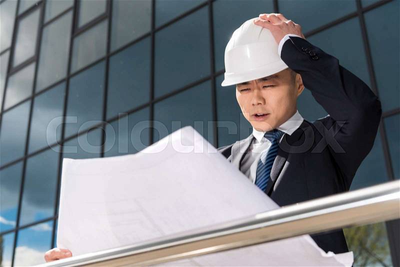 Portrait of confused professional architect in hard hat looking at blueprint, stock photo