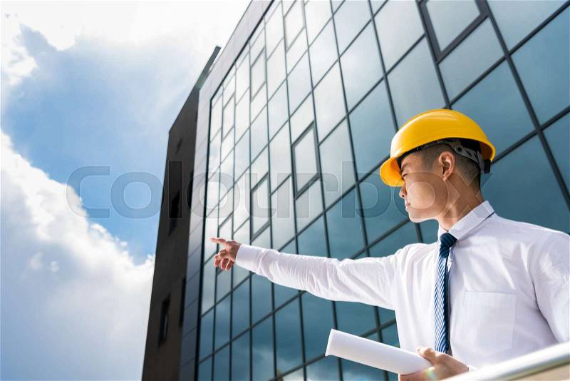 Side view of professional architect in hard hat holding blueprint and gesturing, stock photo