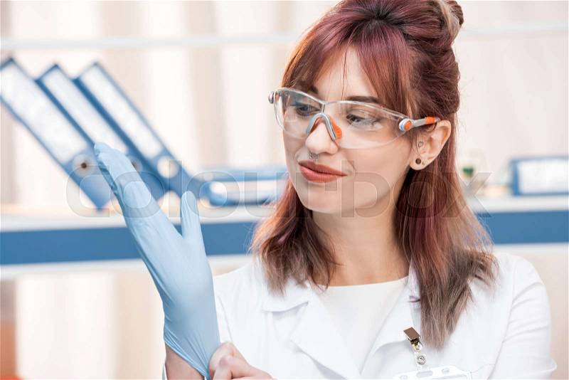 Young scientist in lab coat goggles wearing medical glove in chemical lab, stock photo
