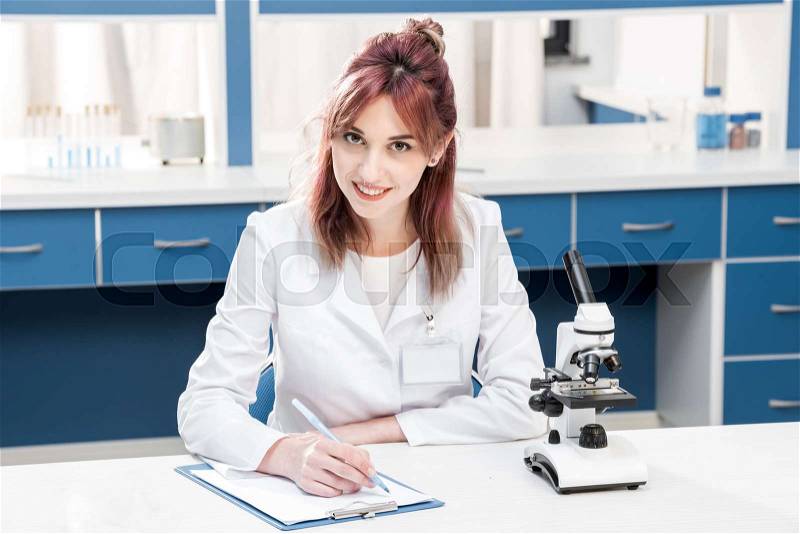 Young scientist in lab coat writing in folder and looking at camera in chemical lab , stock photo