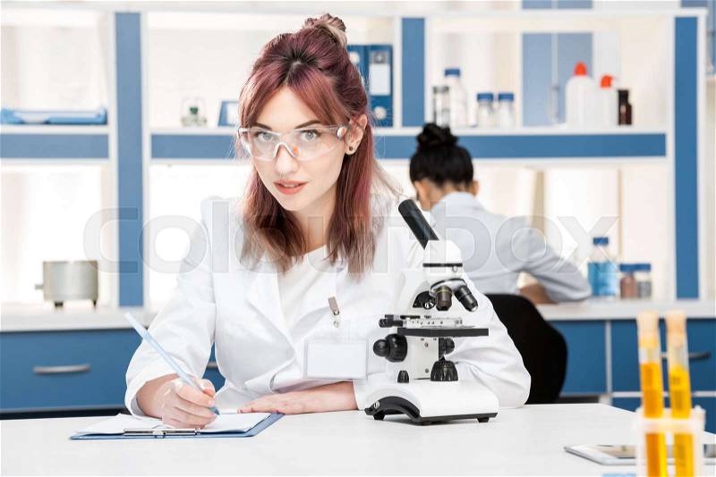 Young scientist in lab coat working with microscope and writing in folder in chemical lab , stock photo