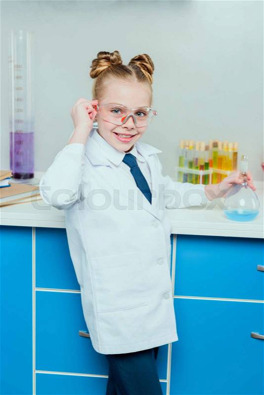 Schoolgirl in protective goggles and white coat holding flask with reagent in science laboratory, stock photo