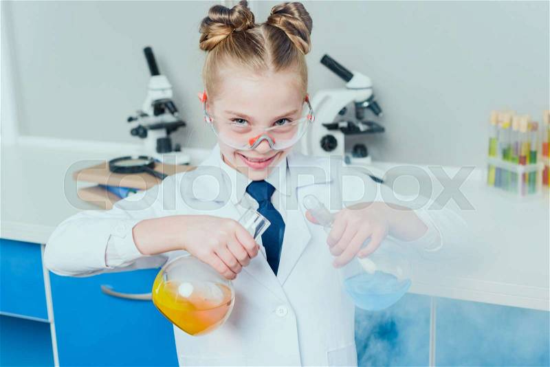 Little scientist in lab coat and protective eyeglasses making experiment with reagents in flasks in science laboratory, stock photo