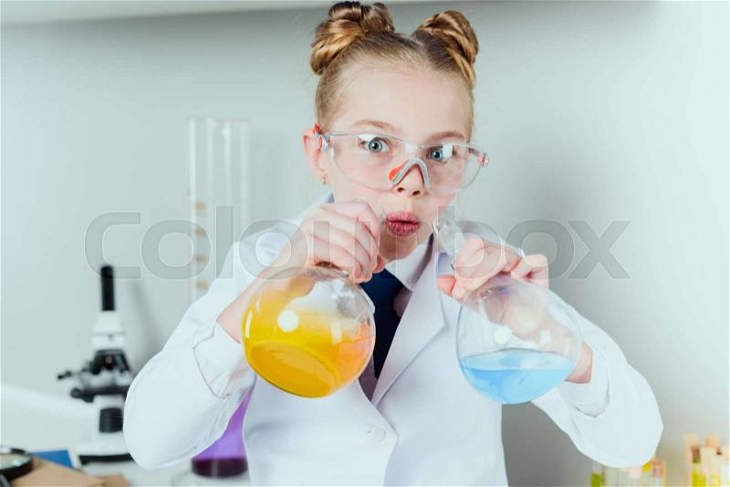 Little scientist in lab coat and protective eyeglasses making experiment with reagents in flasks in science laboratory, stock photo
