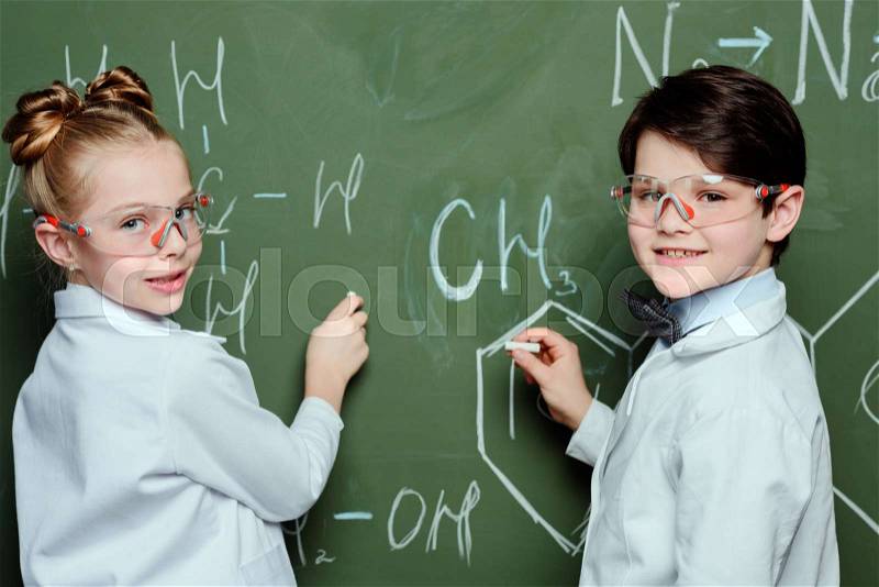 Schoolchildren in white coats and protective eyeglasses drawing chemical formulas on chalkboard and looking at camera, science school concept, stock photo