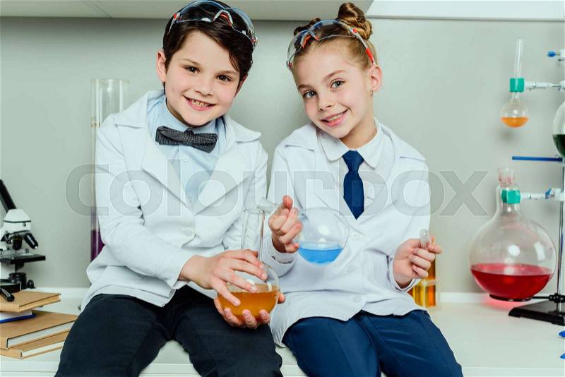 Schoolchildren in white coats holding reagents in flasks sitting in science laboratory, stock photo