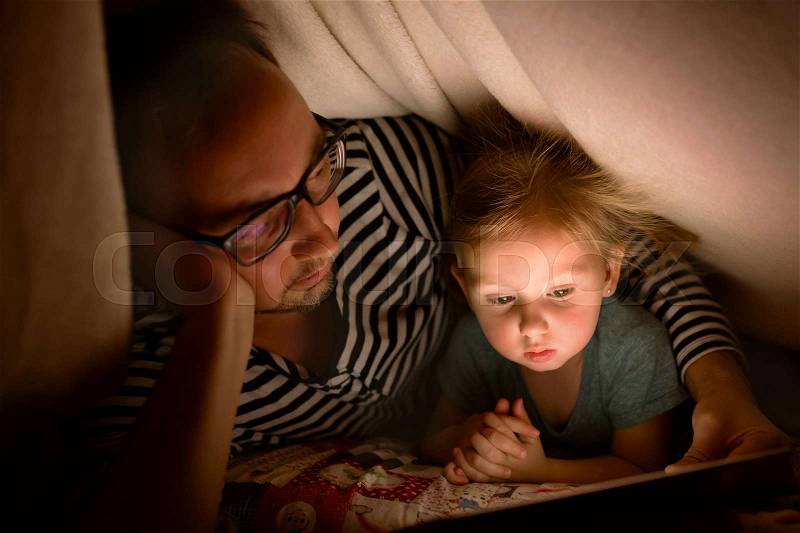 Young father at home at night with his cute little daughter playing or watching something on tablet, stock photo