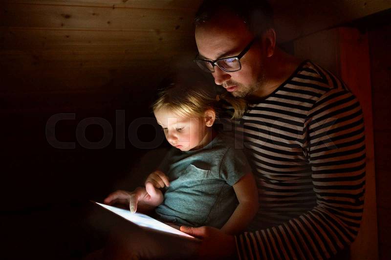 Young father at home at night with his cute little daughter on his lap playing or watching something on tablet, stock photo