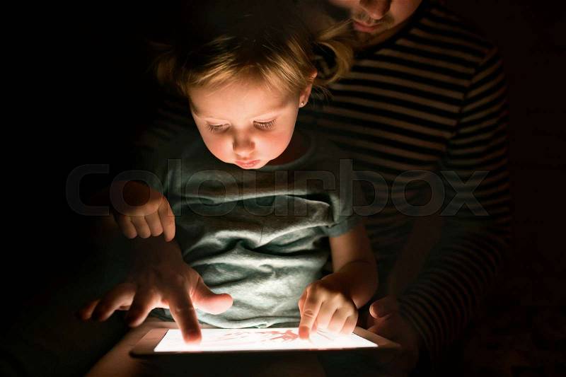 Unrecognizable father at home at night with his cute little daughter on his lap playing or watching something on tablet, stock photo