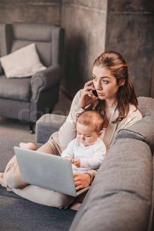 Young mother combine of child\'s care and remote working at home. Woman talking on smartphone and using laptop with her little baby boy, stock photo