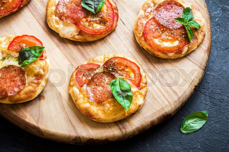 Mini Pizzas - Fresh homemade mini pizzas with pepperoni, cheese, tomatoes and basil on rustic black stone background with copy space, stock photo
