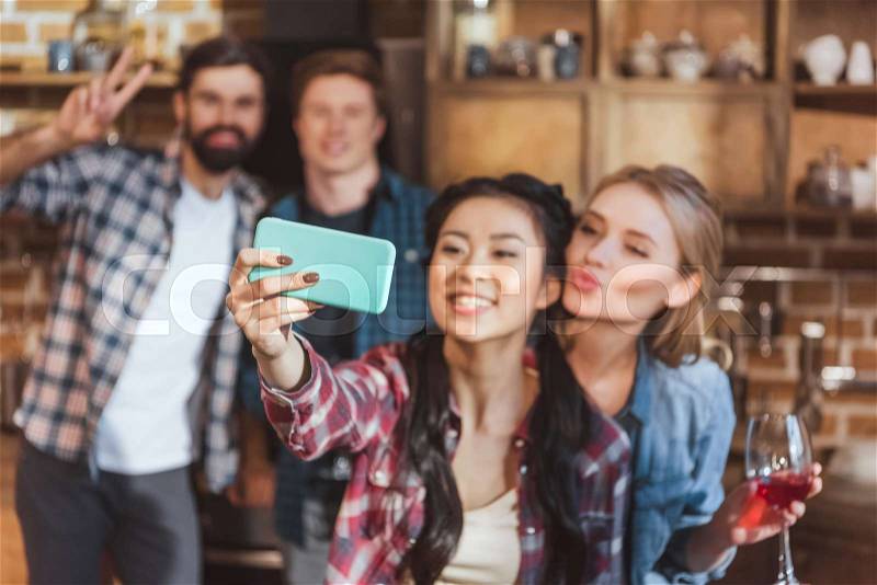 Young people at home party. Girls taking selfie with boys standing blurred on background, stock photo
