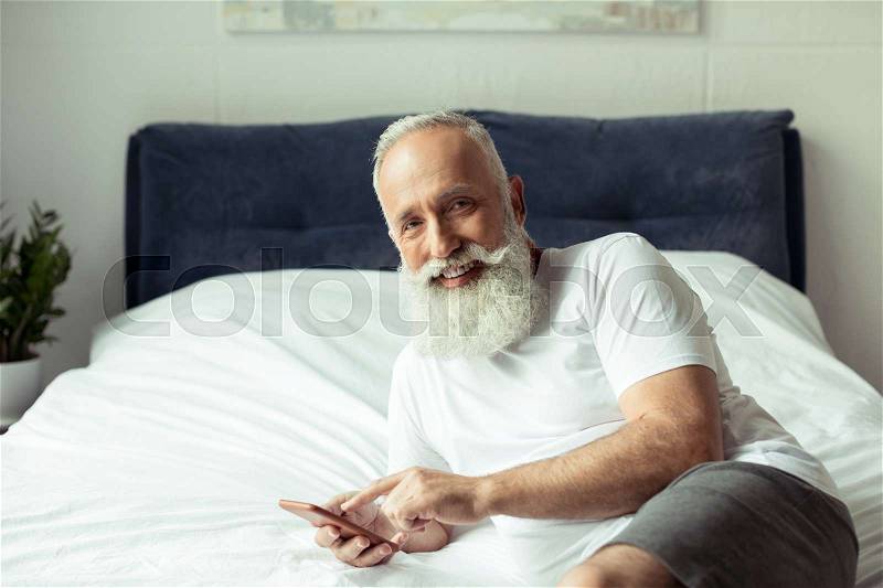 Bearded senior man using smartphone while lying on bed, stock photo