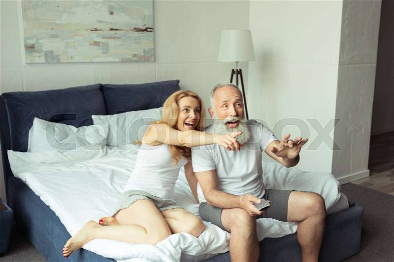 Happy mature couple laughing and pointing on tv while sitting on bed at home, stock photo