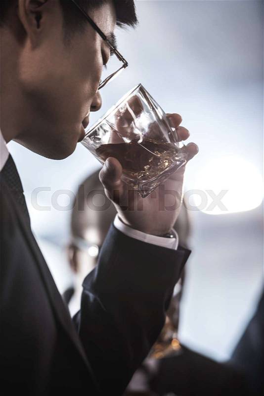 Close-up view of asian businessman in eyeglasses drinking whiskey from glass, stock photo