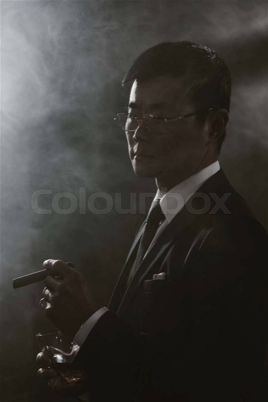 Portrait of confident asian businessman in suit with glass of whiskey smoking cigar , stock photo