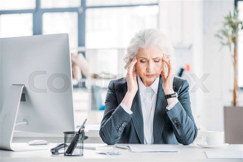 Senior businesswoman with headache sitting at workplace with desktop computer, stock photo