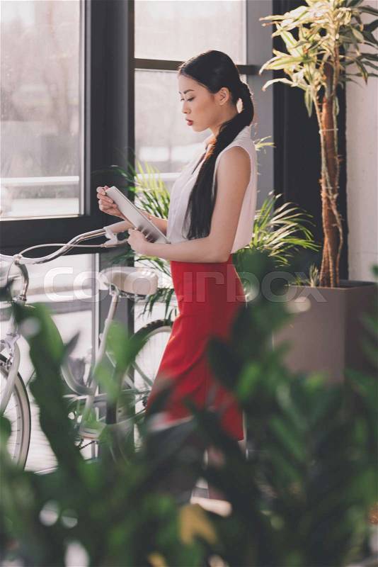 Attractive asian businesswoman working with digital tablet in office, bicycle standing behind, stock photo