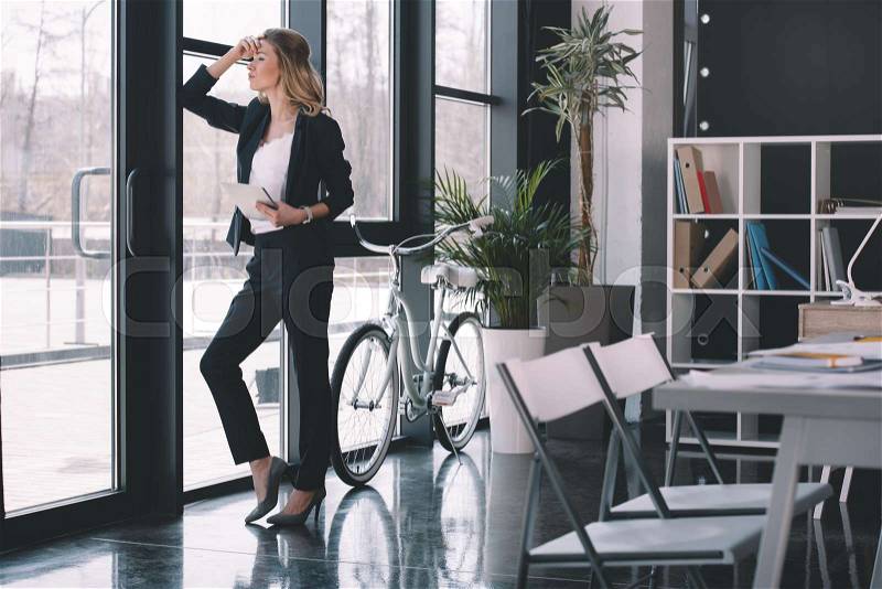 Attractive pensive businesswoman in suit with digital tablet standing at window in office, bicycle standing behind, stock photo