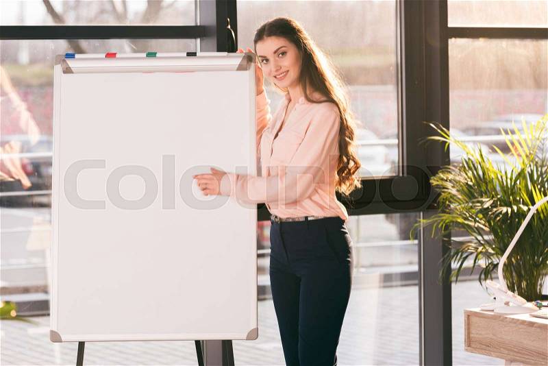 Smiling young businesswoman making presentation in blank whiteboard and looking at camera, stock photo