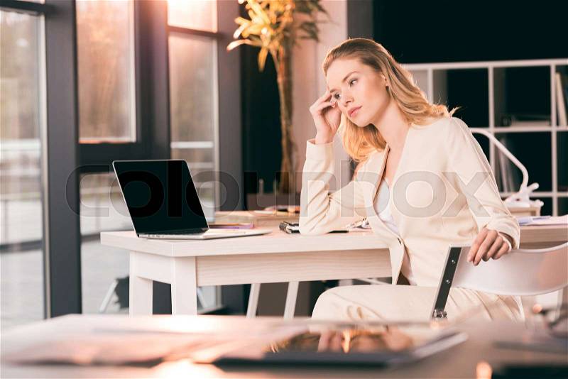 Young businesswoman in suit sitting at table in office and looking aside, stock photo