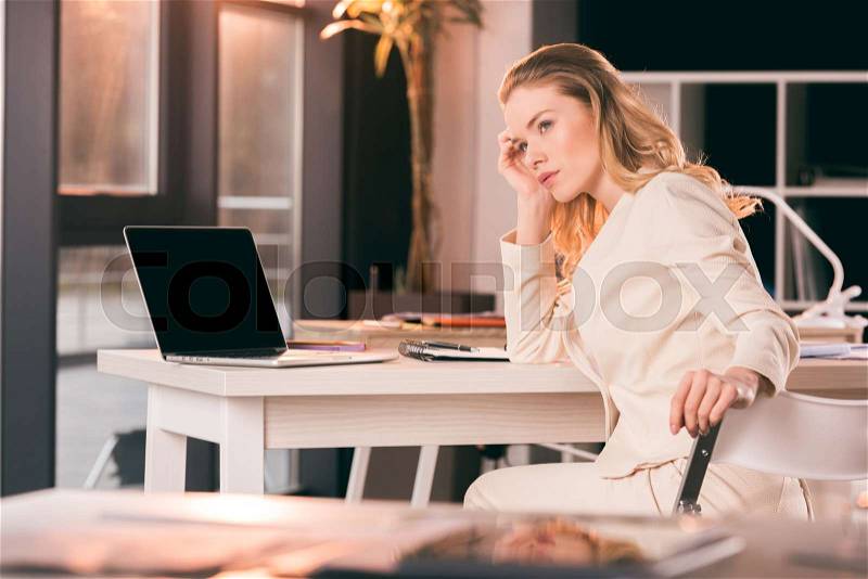 Young thoughtful businesswoman in suit sitting at table in office, stock photo