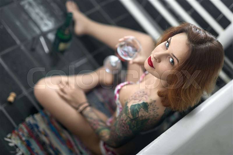 Beautiful girl with colorful tattoos sits near white bath on the motley carpet on the dark tiled floor in the bathroom. She holds a glass and looks into the camera. Top view photo. Indoors, stock photo
