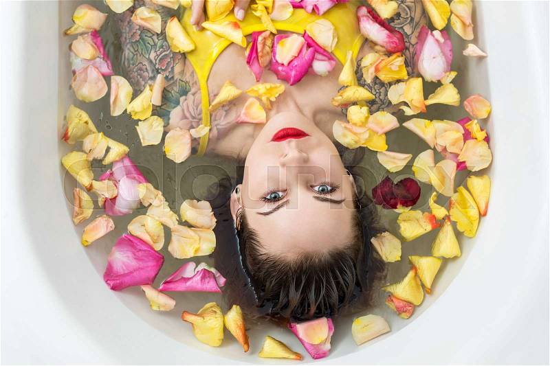 Beautiful wet girl with colorful tattoos lies in the white bath full of water with flower petals. She wears a yellow swimsuit and looks into the camera. Top view photo. Indoors. Horizontal, stock photo