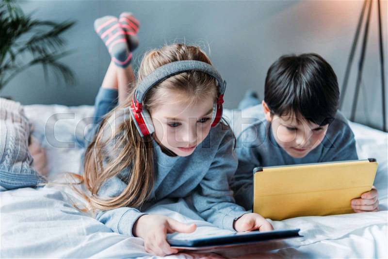 Little girl in headphones and boy using digital tablets while lying on bed, stock photo