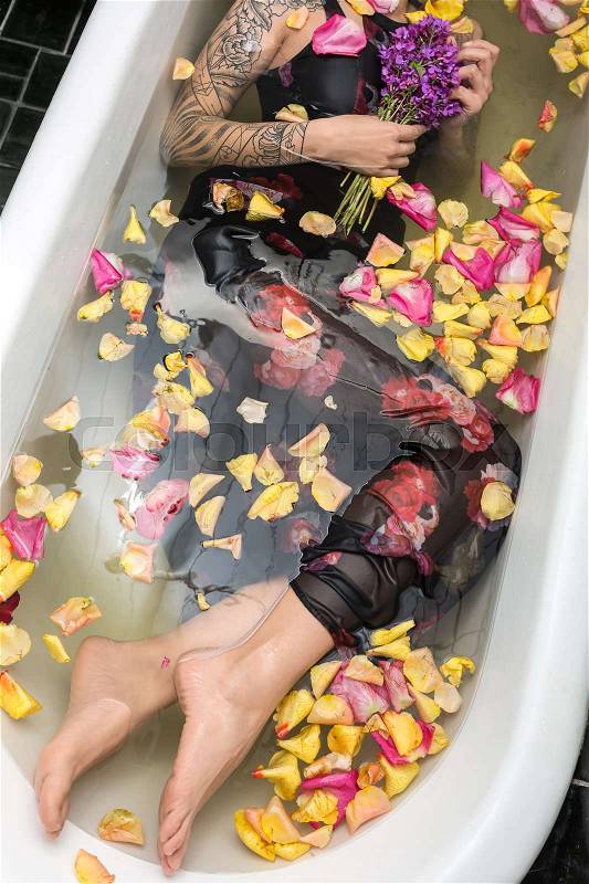 Attractive girl with tattoos lies in the white bath full of water with flower petals. She wears a dark dress with flower prints and holds a violet bouquet in right hand. Top view photo. Indoors, stock photo