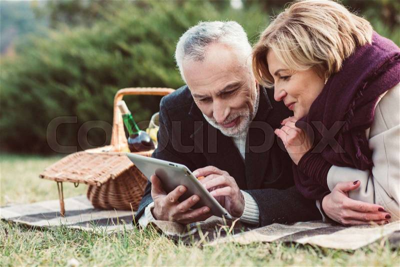 Beautiful smiling mature couple using digital tablet while lying on plaid with picnic basket in autumn park, stock photo