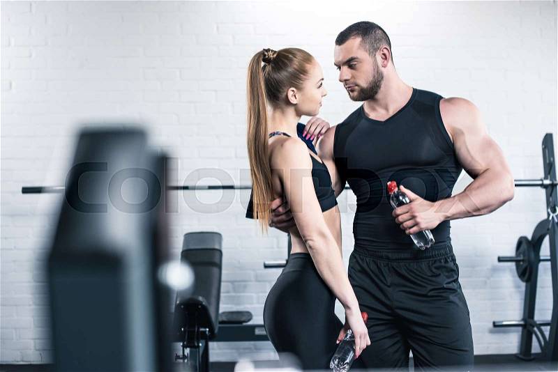 Athletic man in woman holding bottles of water and hugging in gym, stock photo