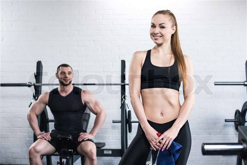 Smiling muscular man looking at sporty woman holding towel and bottle of water in gym , stock photo