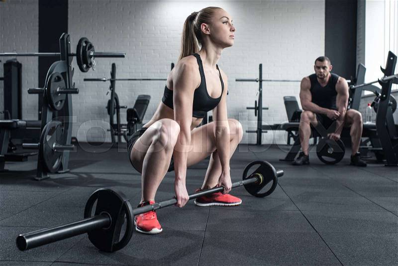 Woman doing strength training with barbell while man sitting at gym, stock photo