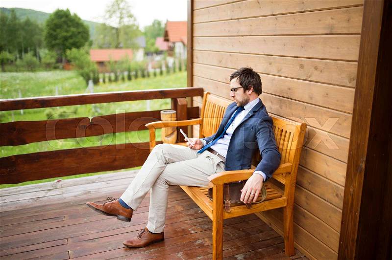 Young businessman at home sitting on front porch, holding smart phone, texting, stock photo