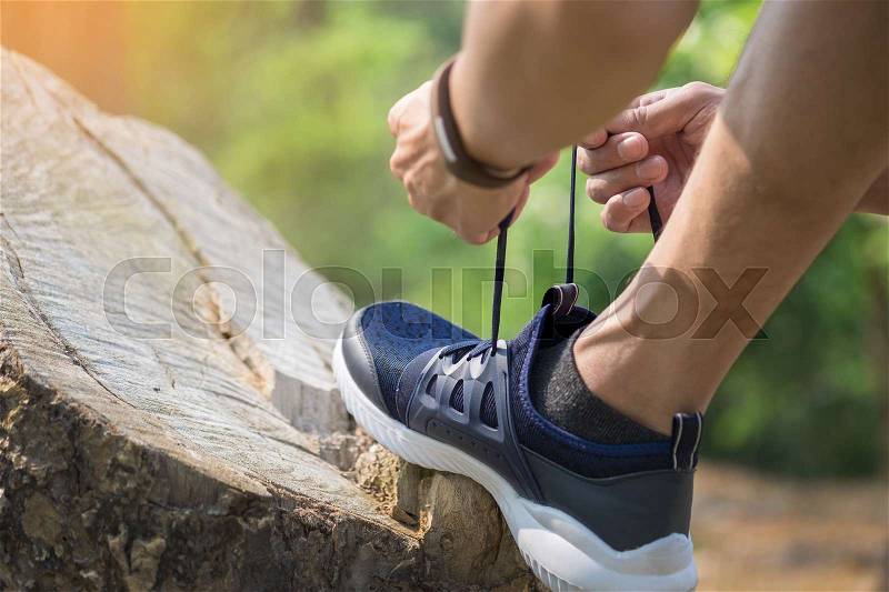 Cropped shot of young man runner tightening running shoe laces, getting ready for jogging exercise outdoors. Male jogger lacing his sneakers standing on forest path before morning run, stock photo