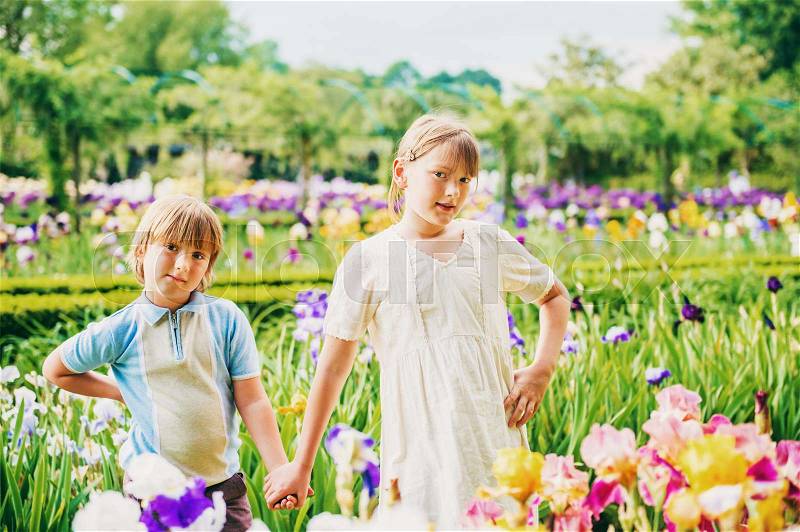 Group of two kids, little boy and girl, posing in beautiful garden, wearing retro style clothing, holding hands. Brother and sister playing together in amazing summer park between iris flowers, stock photo