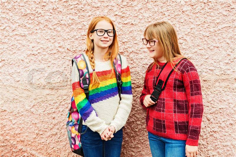 Group of two adorable kid girls posing outdoors against pink wall, wearing glasses, school backpacks and bright colorful pullovers, back to school concept, stock photo