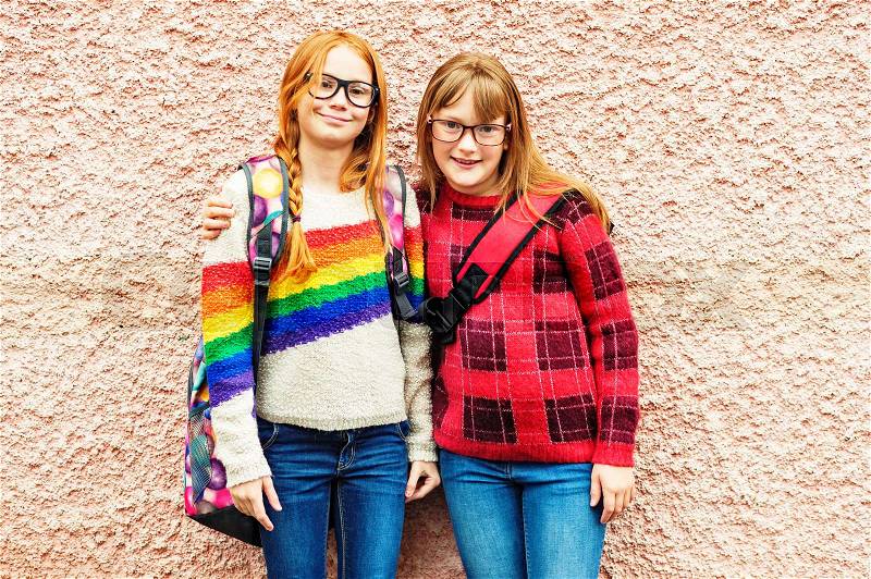 Group of two adorable kid girls posing outdoors against pink wall, wearing glasses, school backpacks and bright colorful pullovers, back to school concept, stock photo