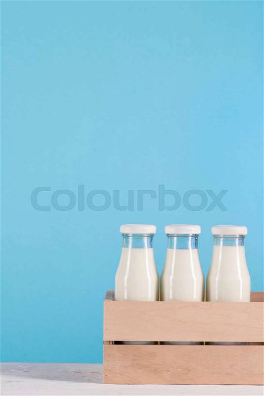 Glass bottles with milk in wooden box on tabletop at blue background, stock photo