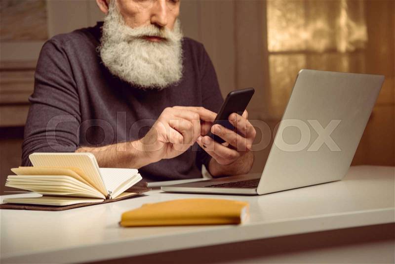 Cropped shot of grey haired man sitting at table and using smartphone at home, stock photo