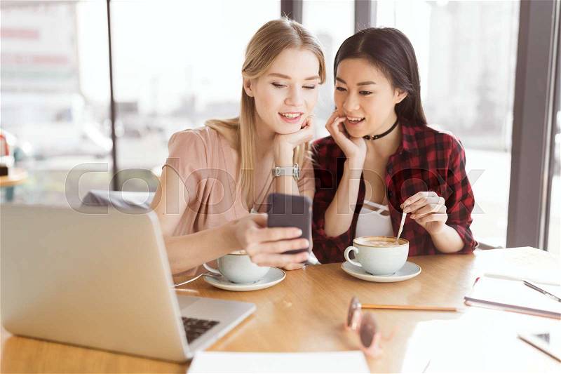 Smiling young women using smartphone while drinking coffee together at lunch meeting, stock photo