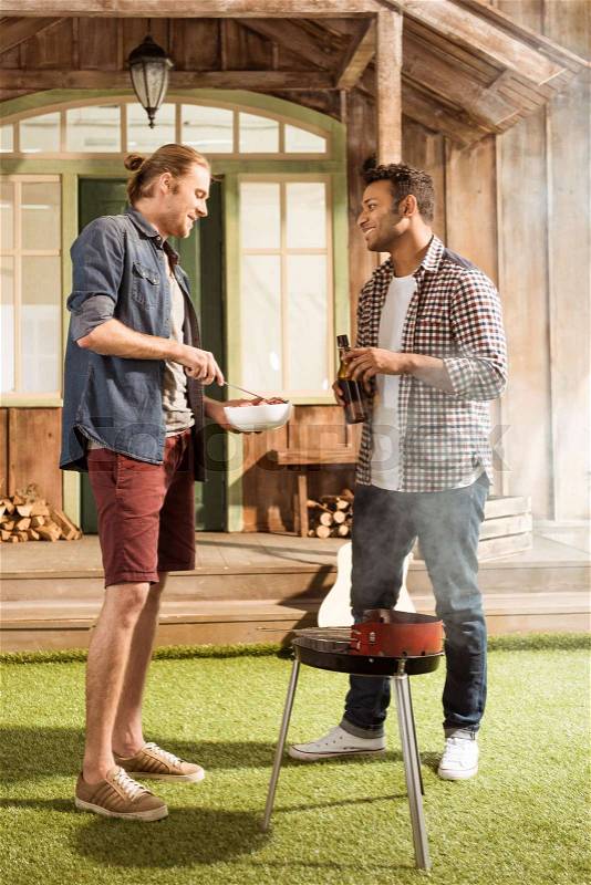 Happy multiethnic men grilling burgers while talking and drinking beer, stock photo