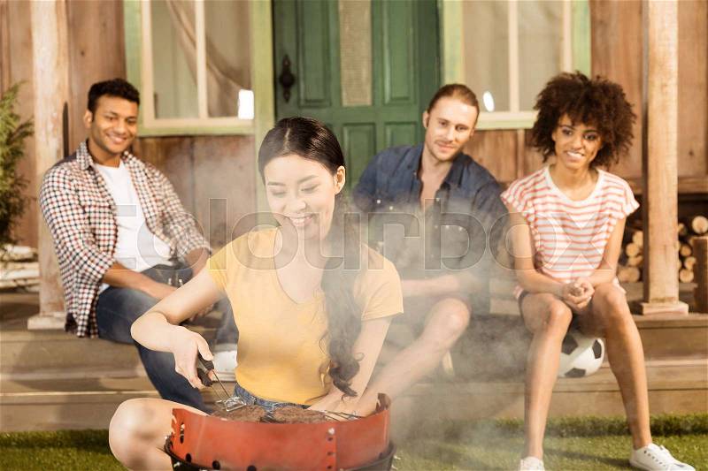 Young woman preparing barbecue grill on patio while her friends sitting at porch, stock photo